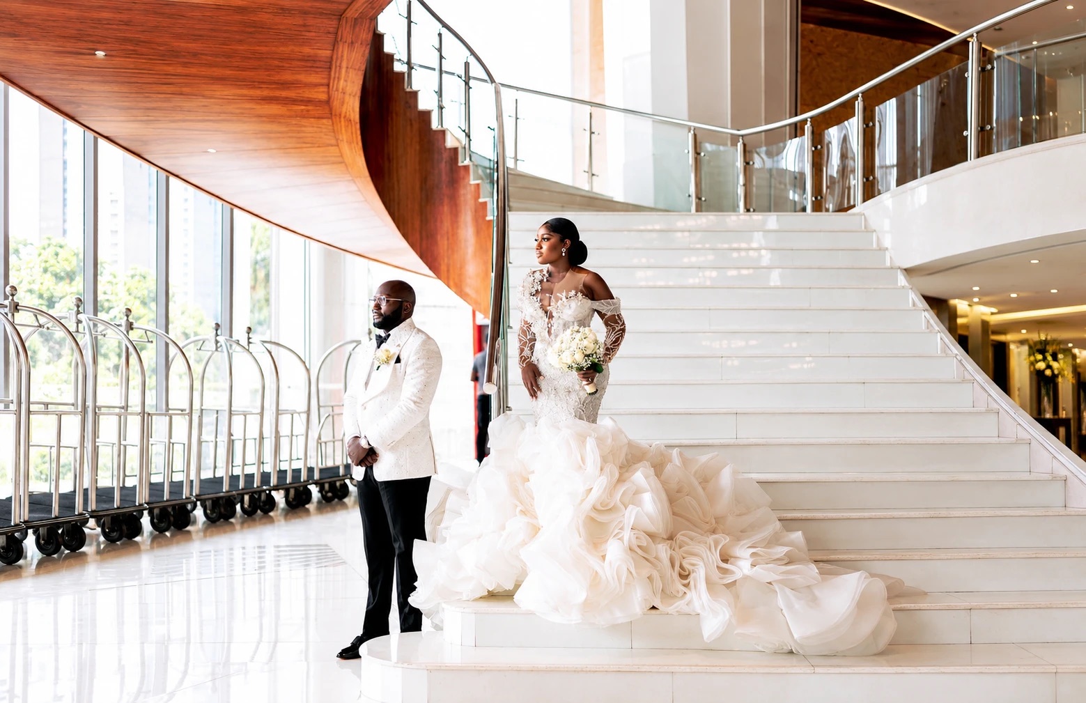 A bride and groom in a bright hotel lobby: the bride on a white staircase in a ruffled gown with a sweeping train, the groom in a white dinner jacket